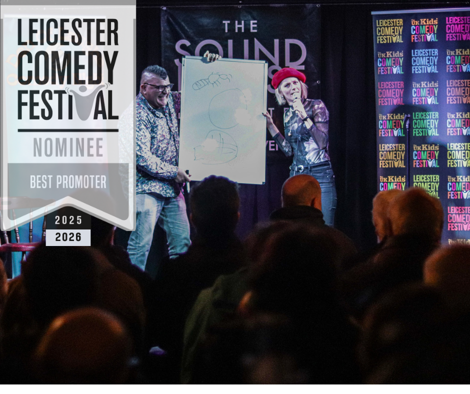 A female comedian is on stage, wearing a red beret and pointing at a whiteboard. An audience member is onstage with her holding the whiteboard. He is laughing. The audience are watching. In the background is the Leicester Comedy Festival banner, and there's a Leicester Comedy Festival Nominee - Best Promoter 2025, 2026 overlay, in black, grey and white.